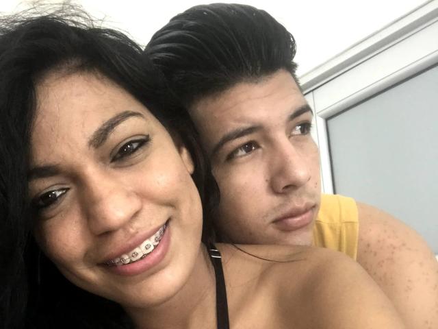 LaSantaKaren with black hair and white teeth smiling at the camera, her arms around a guy with dark hair and a yellow shirt, his face looking bored.
