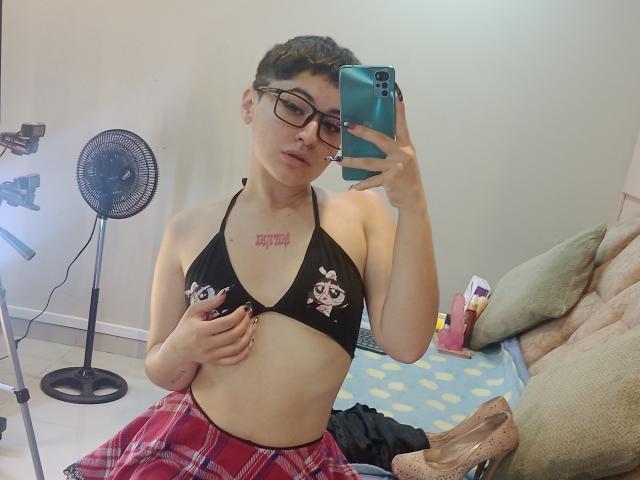 MissTat with short dark hair, glasses, and a black bikini top showing small tits with cat prints, holding a phone, wearing red plaid pants, in a mirror selfie.