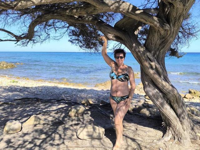 DoraBlueEyess staat onder een kronkelende boom, gekleed in een groene en zwarte bikini met bloemenprint, waarbij haar borsten zichtbaar zijn, met één arm omhoog, en op de achtergrond een zonnig, rotsachtig strand..