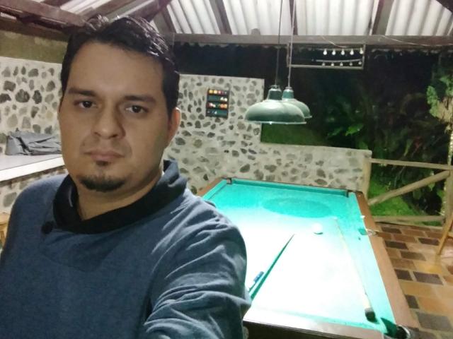 JimmySamuel, a chubby dude with short black hair and a goatee, wearing a blue hoodie, standing in front of a green-lit pool table, taking a selfie.