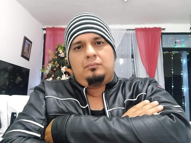JimmySamuel, un homme à la peau mate, portant un bonnet noir à rayures, une veste en cuir noir, les bras croisés, une petite barbe de chèvre, un léger sourire, un sapin de Noël en arrière-plan, des rideaux rouges, une fenêtre avec des guirlandes lumineuses..