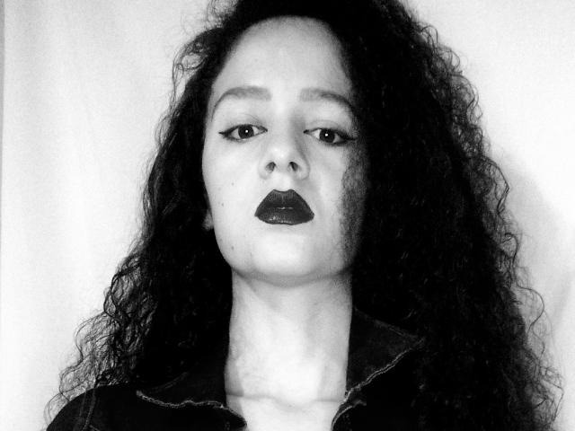Black-and-white photo of Mihiany with curly dark hair, wearing dark lipstick and a black jacket, looking slightly up with a serious expression. Mihiany's nose is centered.