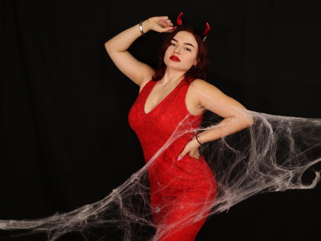NicolPots with red lace dress, devil horns, and web, hand on head, red lipstick, and bracelet, looking at camera, standing against black background, showing her tits.