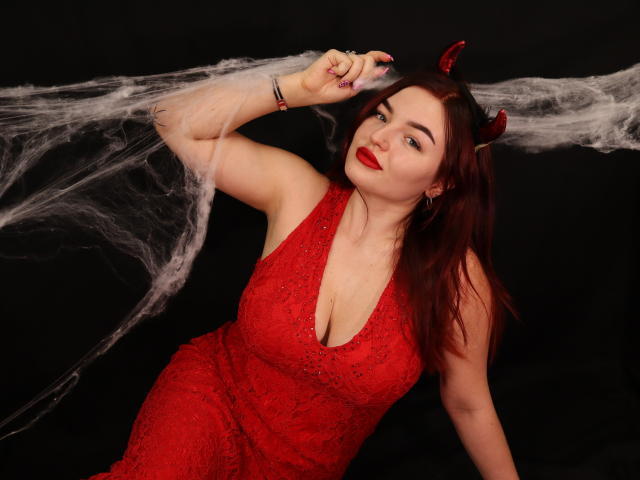 NicolPots with red devil horns, red dress, and red lipstick, showing her tits, with a smoke web in front, against a black background. Her red hair is down.