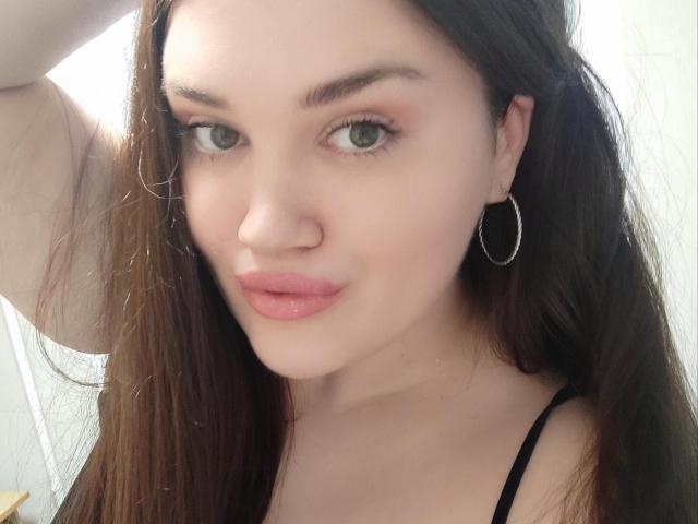 WinterPeggy has pale skin, green eyes, pink lipstick, and brown hair, wearing hoop earrings and a black top, with a slight smile and raised arm.