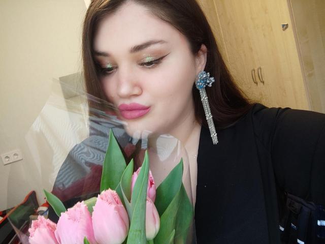 WinterPeggy with pink lipstick, green eyeshadow, and blue earrings, smelling pink tulips through a clear plastic wrap, wearing a black top, brown hair, and fair skin.