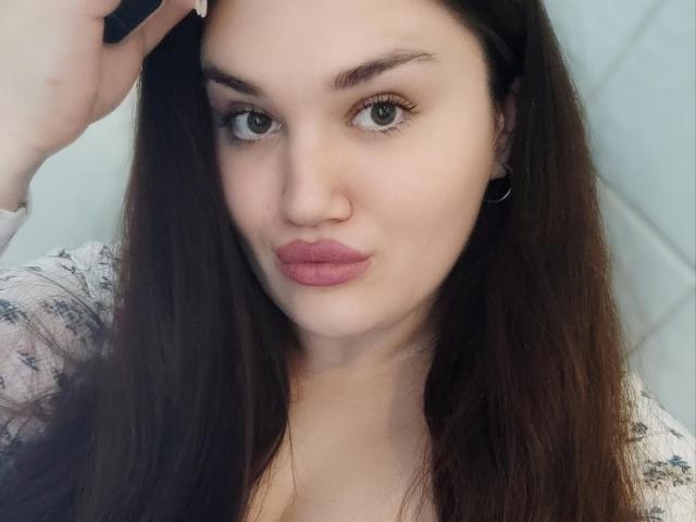 WinterPeggy with long brown hair, full pink lips, and green eyes, holding a phone to her ear, wearing a white patterned shirt, small black earrings.