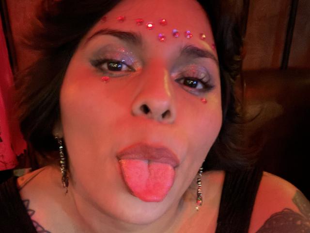 Chili with dark hair, red face paint on forehead, tongue sticking out, red lips, wearing black tank top, earrings.