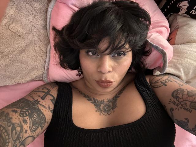 Chili, a tattooed milf with short black hair, lies on pink pillows, wearing a black tank top, looking up.