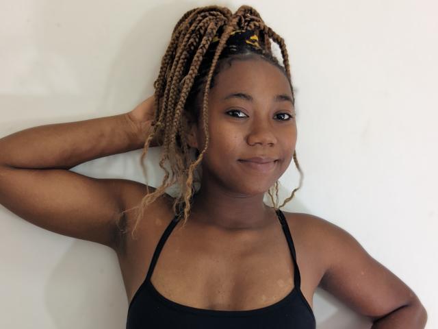 Elyanne with dark brown skin and curly blonde braids, wearing a black spaghetti strap top, has a slight smile, hand in hair, looking at camera.