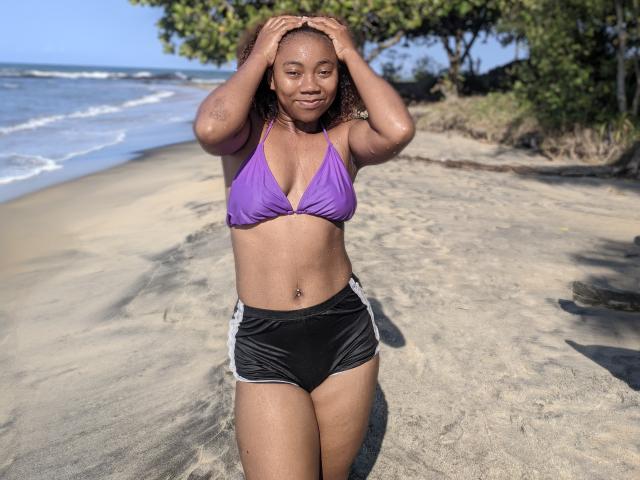 Elyanne with dark brown skin, small purple bikini top showing her tits, black shorts with white stripes, hands in her curly hair, standing on sandy beach, smiling.