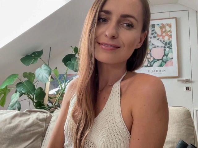VickieBlack, une femme attrayante avec de longs cheveux bruns, portant un haut blanc à encolure dégagée, souriant à la caméra, montrant sa petite poitrine et ses bras fins, assise sur un canapé beige..