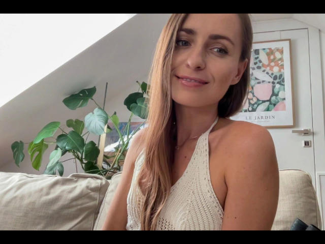 VickieBlack-hot has long brown hair, small tits, wearing a white knitted halter top, smiling at the camera, with green plants and a colorful wall art in the background.