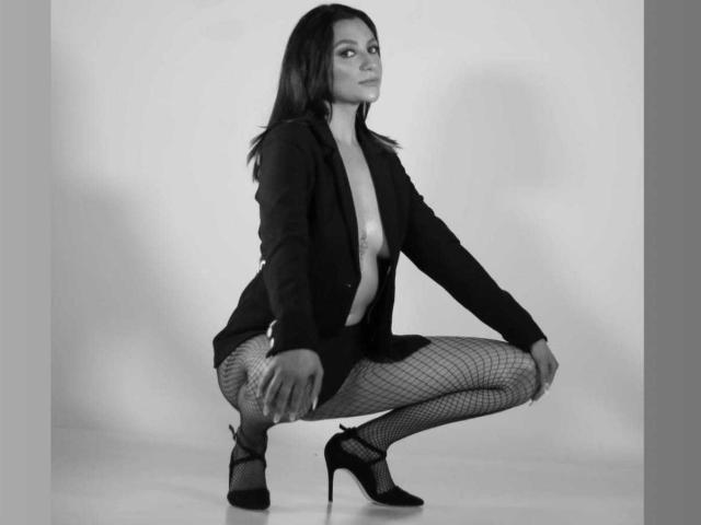 Alizep, squatting, in black blazer, fishnet stockings, and high heels, with her tits exposed, has dark hair and a confident look, in a black-and-white photo.