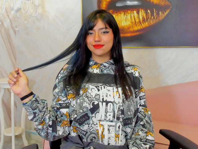 SussiieConor with long black hair, wearing a grey and white patterned shirt, holding her hair, red lipstick, and a black bracelet, sitting on a pink chair.
