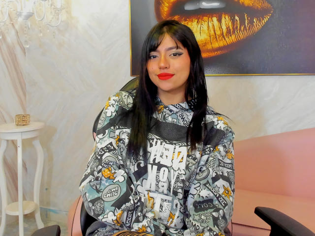 SussiieConor with black hair, red lipstick, and a gray patterned hoodie, sitting on a pink chair, smiling, wearing a black mesh top, looking cute and sexy.