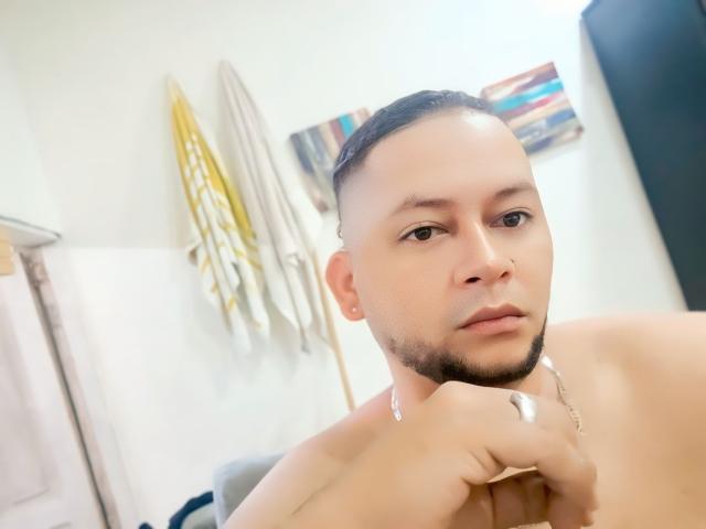 Rengih has a shaved head, a thin beard, and a neutral expression. He's shirtless with a necklace and a ring on his right hand. His face is centered.