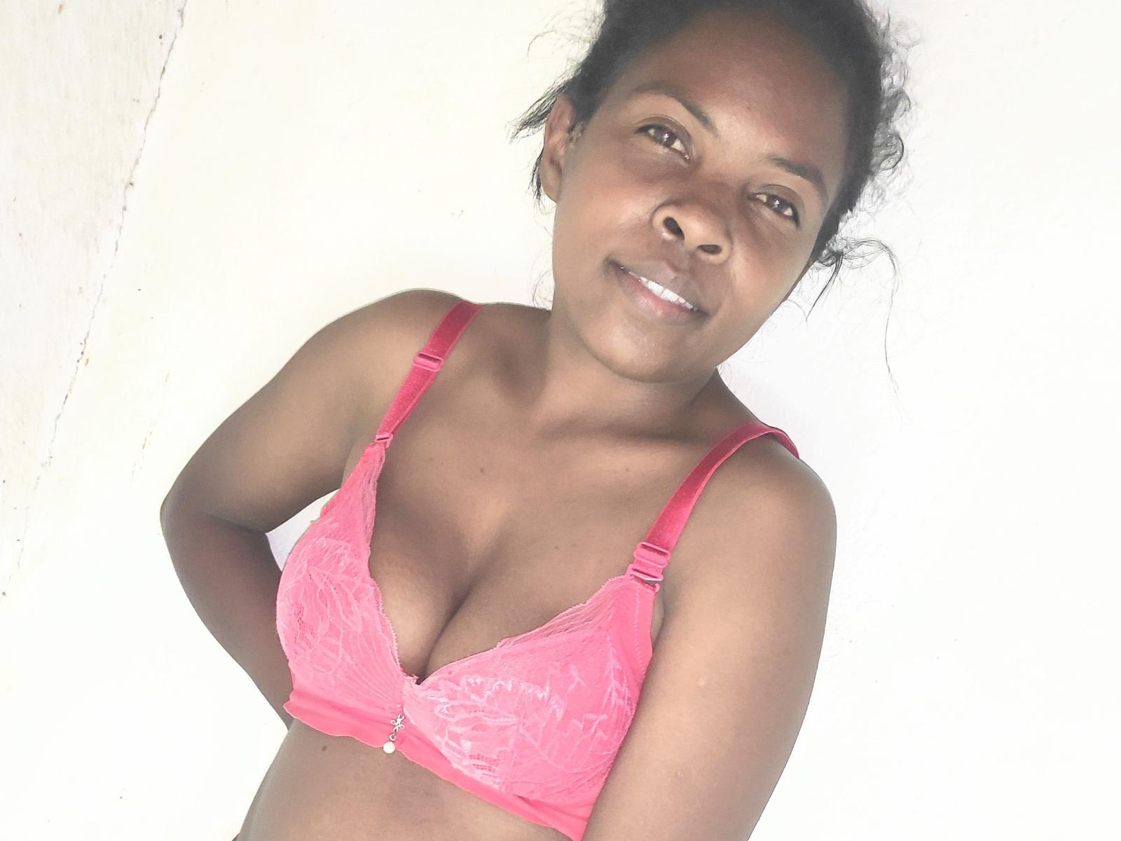 Hobii with dark skin, curly black hair, and small tits wearing a pink lace bra, smiling at the camera. Her hands are on her hips.