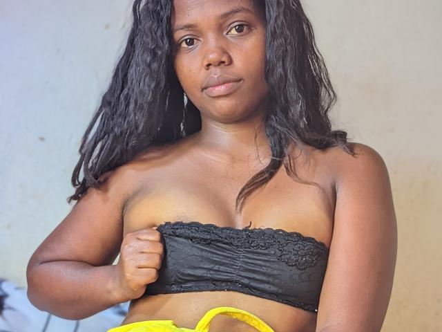 Stechykatte with dark brown skin, wavy black hair, and yellow underwear, pulling down a black lace top to reveal her small, firm tits. She has a serious expression.