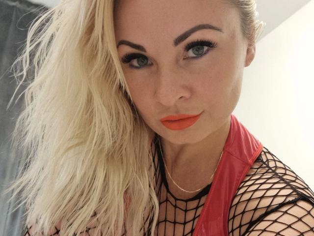 JanePussyCam-hot with blonde hair, blue eyes, orange lipstick, and fishnet top, looking seductively at the camera. Her tits are visible through the net.