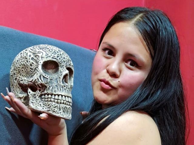 AllizeWalkers with long black hair holding a detailed skull, pouty lips, and a cute expression, against a red background, showing off her small nose and round face.