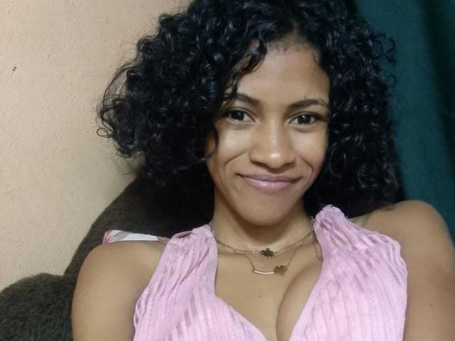 Alya69x with curly black hair, brown skin, smiling, wearing a pink ribbed top showing her tits, and two gold necklaces. Her tits are noticeable and she's cute.