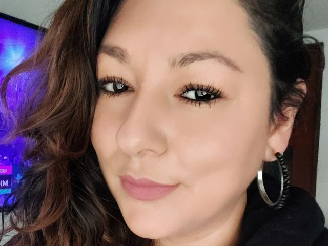 SakotoDika's face, with wavy brown hair, hoop earrings, and pink lipstick, smiling slightly, showing dark eyelashes and well-groomed eyebrows, in a selfie.