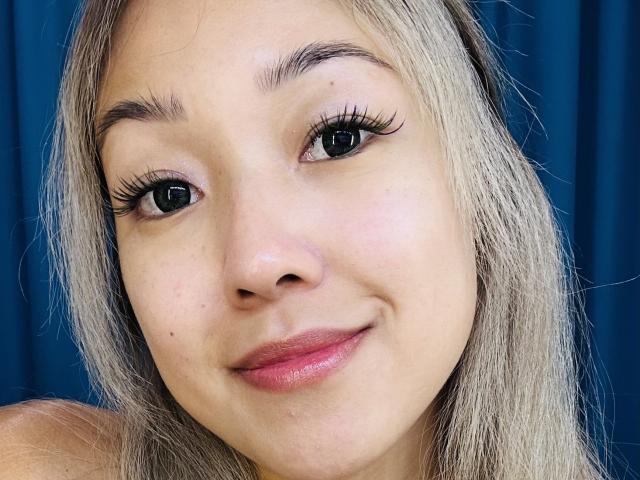 RenyLime's face, showing light skin, blonde hair, black eyelashes, small nose, pink lips, and a slight smile, with a blue background. Boobs not visible, focus on face.