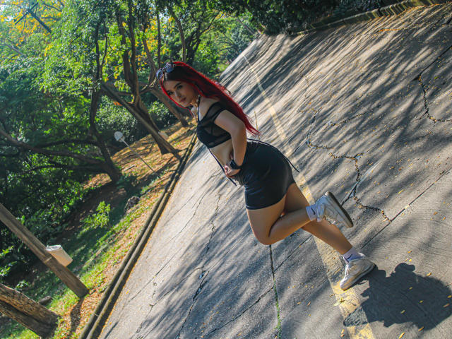 Leylak, con cabello rojo, un top corto negro y una minifalda negra que deja ver su trasero, de pie en una acera agrietada, con zapatillas blancas, mirando hacia atrás, en un parque soleado..