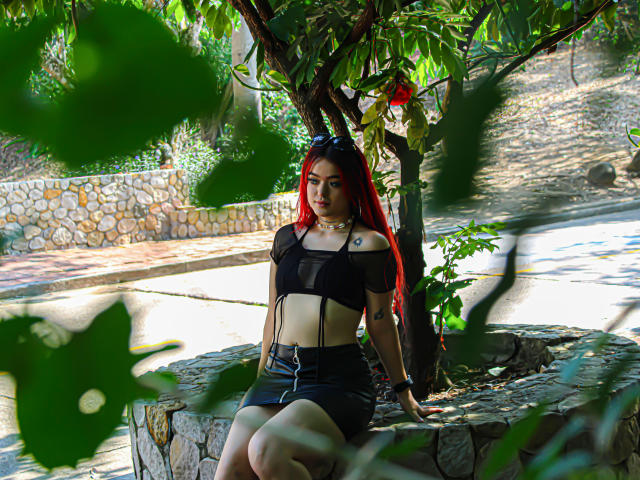 Leylak, avec ses cheveux roux, son haut en maille noire et sa jupe noire, assise sur une corniche en pierre, regardant en bas, sous un arbre aux feuilles, portant des lunettes de soleil noires, avec un petit tatouage sur l'épaule droite..