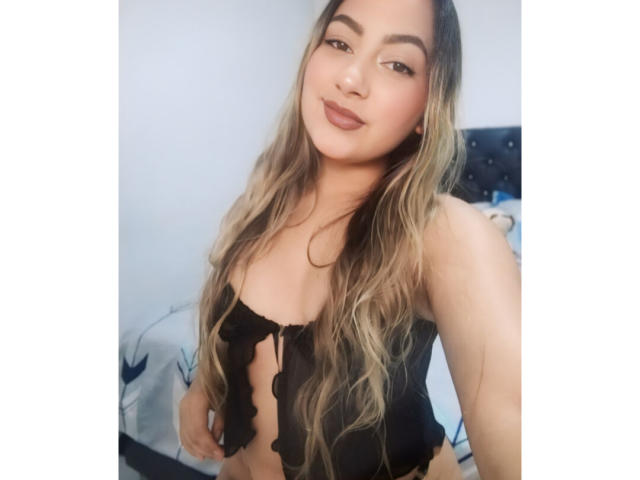 CamilaTeffey with long, wavy blonde hair, wearing a black, ruffled, strapless top, showing her small tits and smiling softly at the camera.