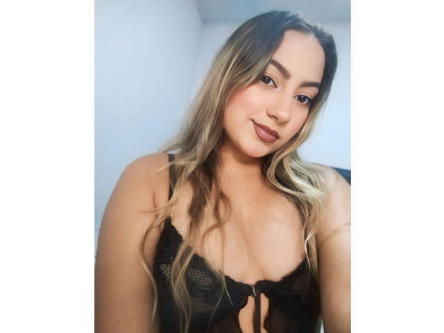 CamilaTeffey with long blonde hair, wearing a black lace bra, showing her small tits, smiling slightly, with soft makeup and a cute face, in a bright photo.