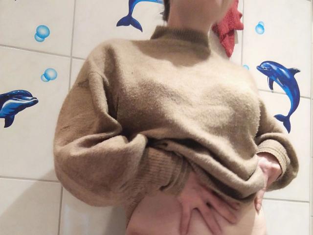 Jennylove-hot lifting a brown sweater to reveal her bare belly and hands resting on her lower stomach, with blue dolphin decals on the tiled wall behind her.