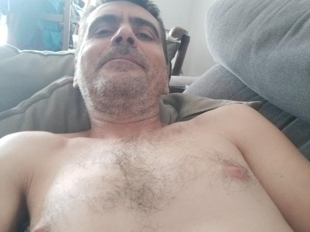 Deifmell, um homem de meia-idade com peito peludo e barba, está deitado sem camisa em um sofá, com seus pequenos mamilos rosados visíveis, e exibe um sorriso leve e confiante..