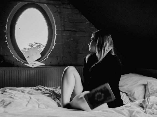 HollyCandyX is sitting on a bed with her legs up, wearing a dark shirt, holding a book, looking out a circular window, in a black-and-white photo.