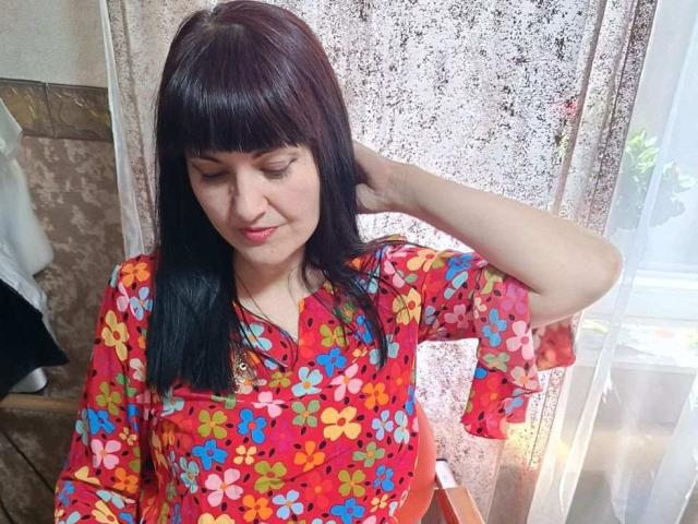 YanaFiesta, com cabelo castanho escuro, vestindo uma camisa vermelha com estampas florais, com a mão no cabelo, seios pequenos, lábios rosados, em pé perto de uma cortina de renda, sorrindo levemente..