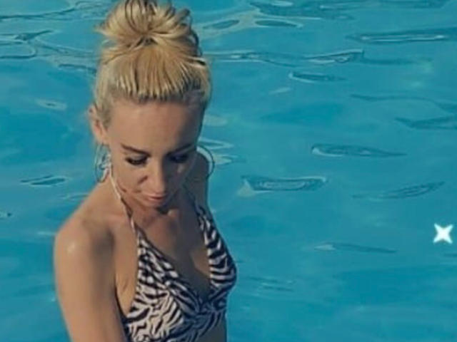 OlixSew a les cheveux blonds attachés en chignon, elle porte un haut de maillot de bain à rayures zébrées, avec de petites seins, et elle se tient dans une piscine bleue, regardant vers le bas avec un léger sourire..
