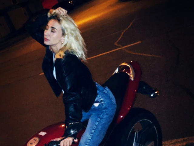 OlixSew with blonde hair, leather jacket, and blue jeans, riding a red motorcycle at night, her ass slightly raised, eyes closed, and hand in her hair.
