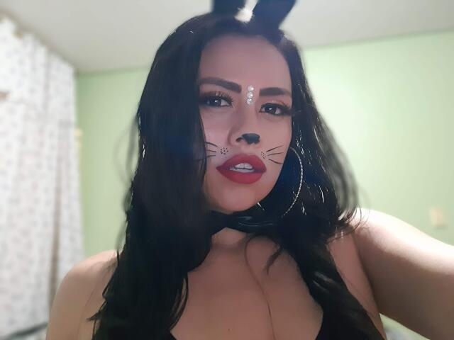 BellaSmithX69 with black hair, cat ears, painted whiskers, red lips, hoop earrings, and face paint, showing her tits in a low-cut top, in a green-walled room.