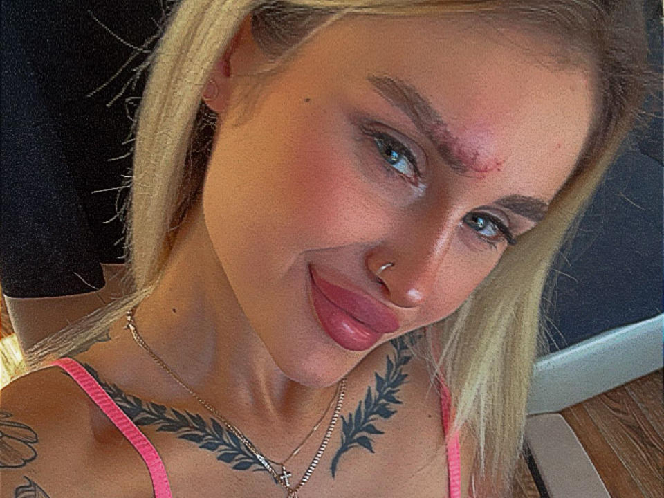 Ravenaa with blonde hair, blue eyes, and a nose piercing, has a red mark on her forehead, tattoos on her chest, and is smiling with her tongue out.