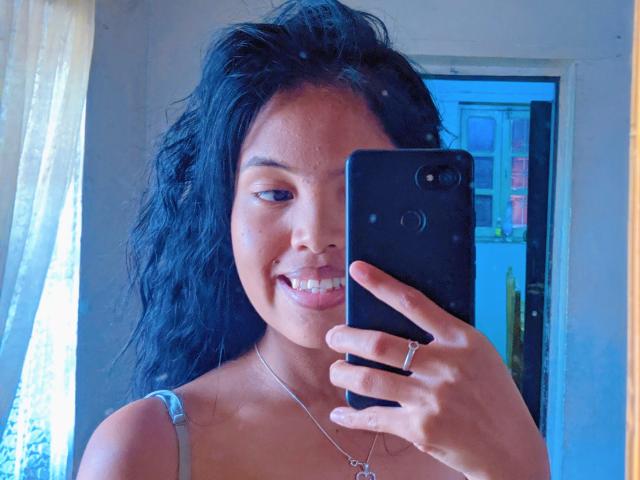 MickyHottieX with curly black hair, smiling, holding a black phone, showing a cute face, wearing a gray tank top, and a silver necklace with a ring.