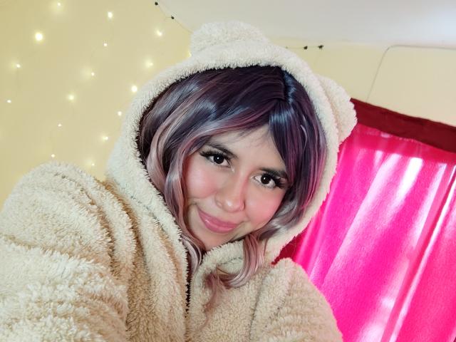MaiaHairy with pink and purple hair, cute smile, fluffy beige bear hoodie, soft skin, and large eyes, standing in front of pink curtain.