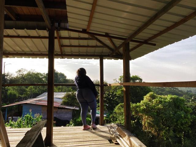 ClarieHaris stands on a wooden porch with a metal roof, facing away, showing her big ass in tight gray leggings and a black top, with pink shoes.
