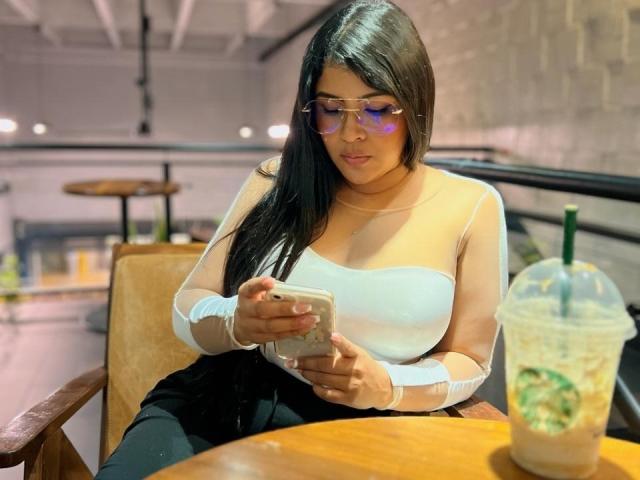 CanelleBom with long black hair, wearing transparent sunglasses, white long-sleeve top, and black pants, looking at phone, with iced drink on wooden table. Boobs visible under top.
