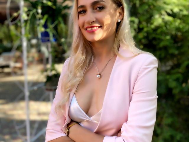 KateOweens with blonde hair, pink blazer, and white top showing her tits, smiling, crossed arms, necklace, and greenery in the background.