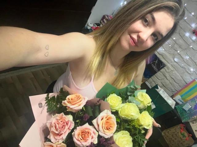 EthelLime with blonde hair, pink tank top, holding bouquet of pink and yellow roses, small heart tattoos on upper arm, smiling at camera, in dimly lit room.
