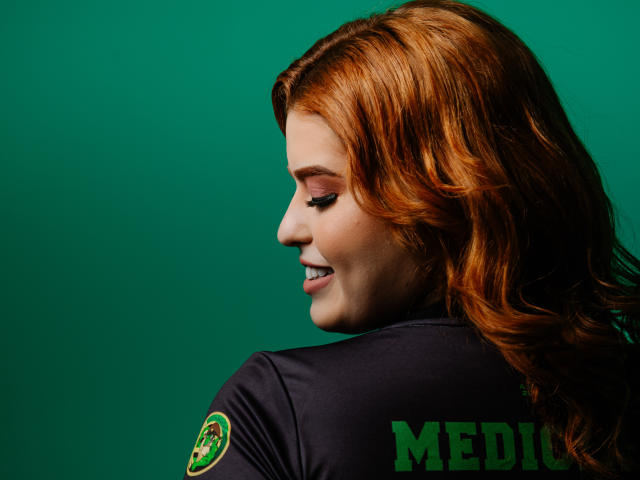 LuanneManu has red, wavy hair and a cute smile, wearing a dark green police uniform with a patch, showing her side profile and sexy ass.