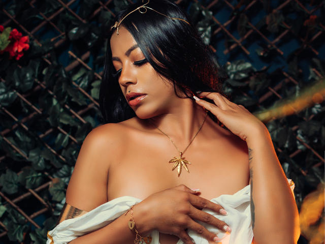 Amberv with dark hair, gold headpiece, and gold necklace, holding a white cloth over her tits, eyes closed, hand touching her neck, against a dark green lattice.