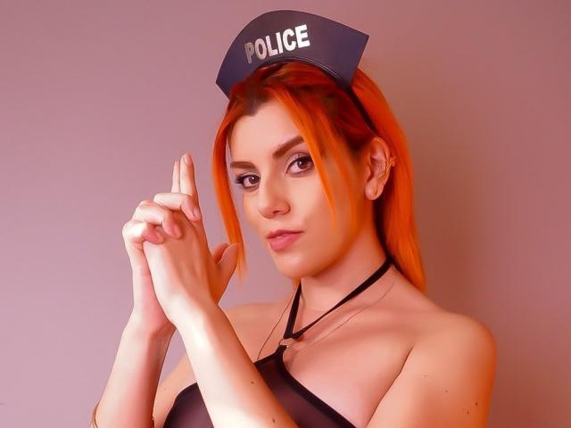 ZoeeMar with bright orange hair, wearing a police hat and black strap top, has her hands together, showing off her tits and a confident, sexy expression.