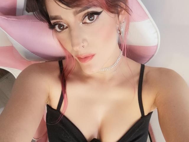 JaneRoa with pink and black hair, black bra, cleavage, pink and white chair, eyeliner, mascara, nose ring, choker, looking at camera, fair skin, soft pink lipstick.